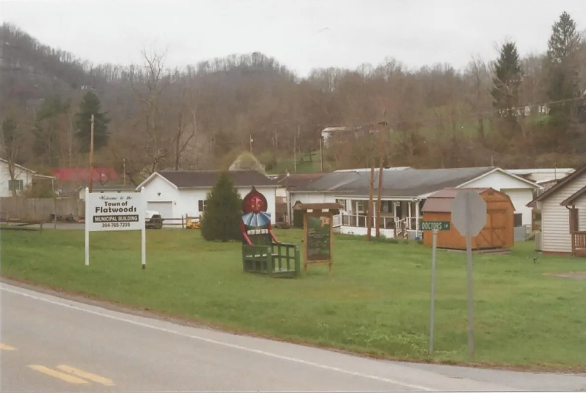 A small-town lawn with a green wooden chair topped by a red-faced, hooded monster figure, beside a town sign, with houses and wooded hills behind