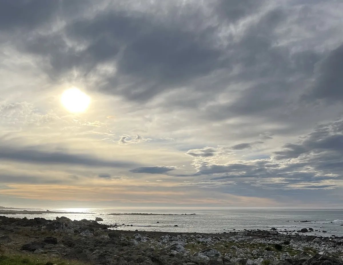 A rocky shoreline and calm sea under a cloudy sky with the sun low behind thin cloud