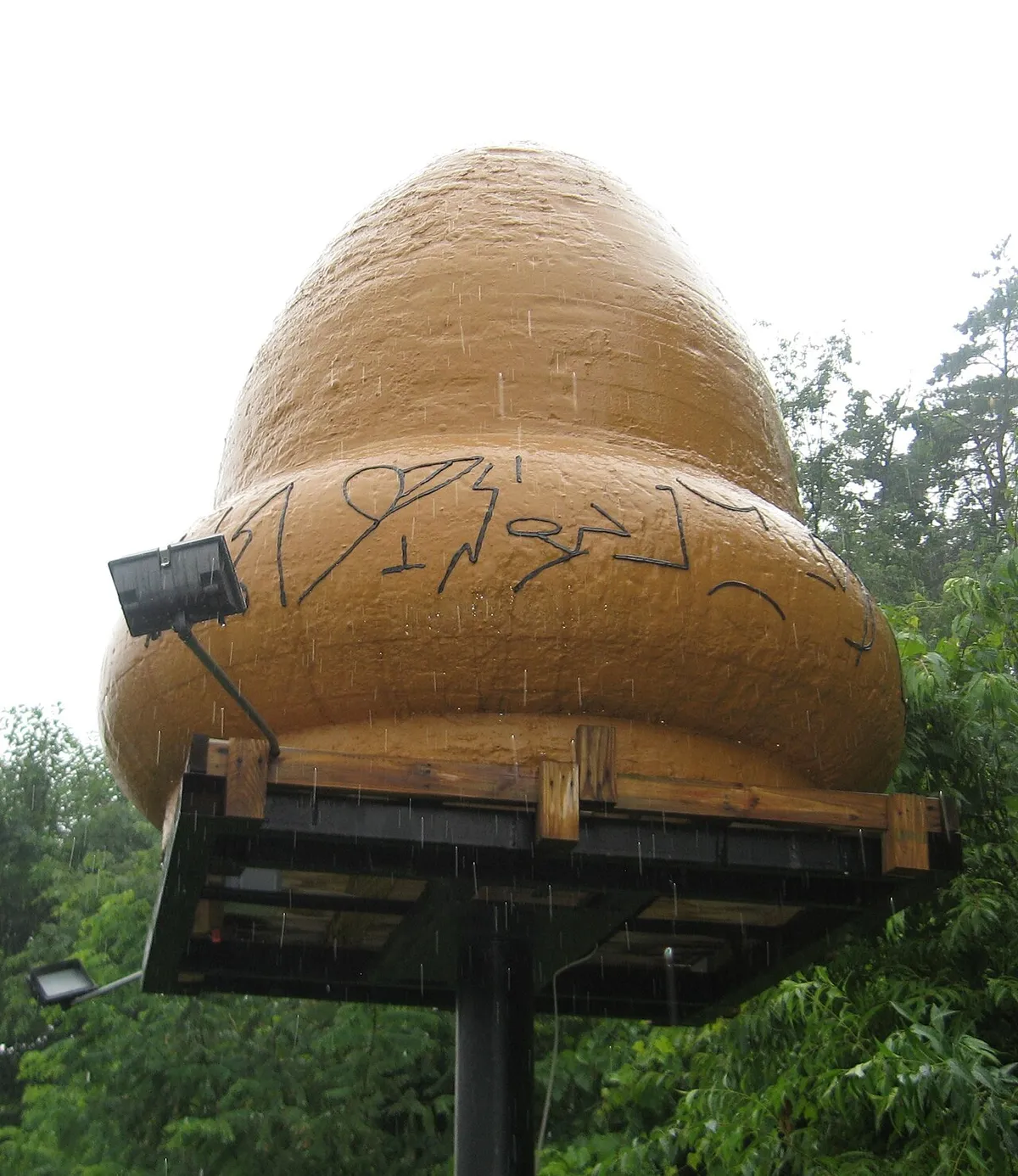 A tan, acorn-shaped model with dark hieroglyph-like marks around its base, on a wooden platform on a pole among trees