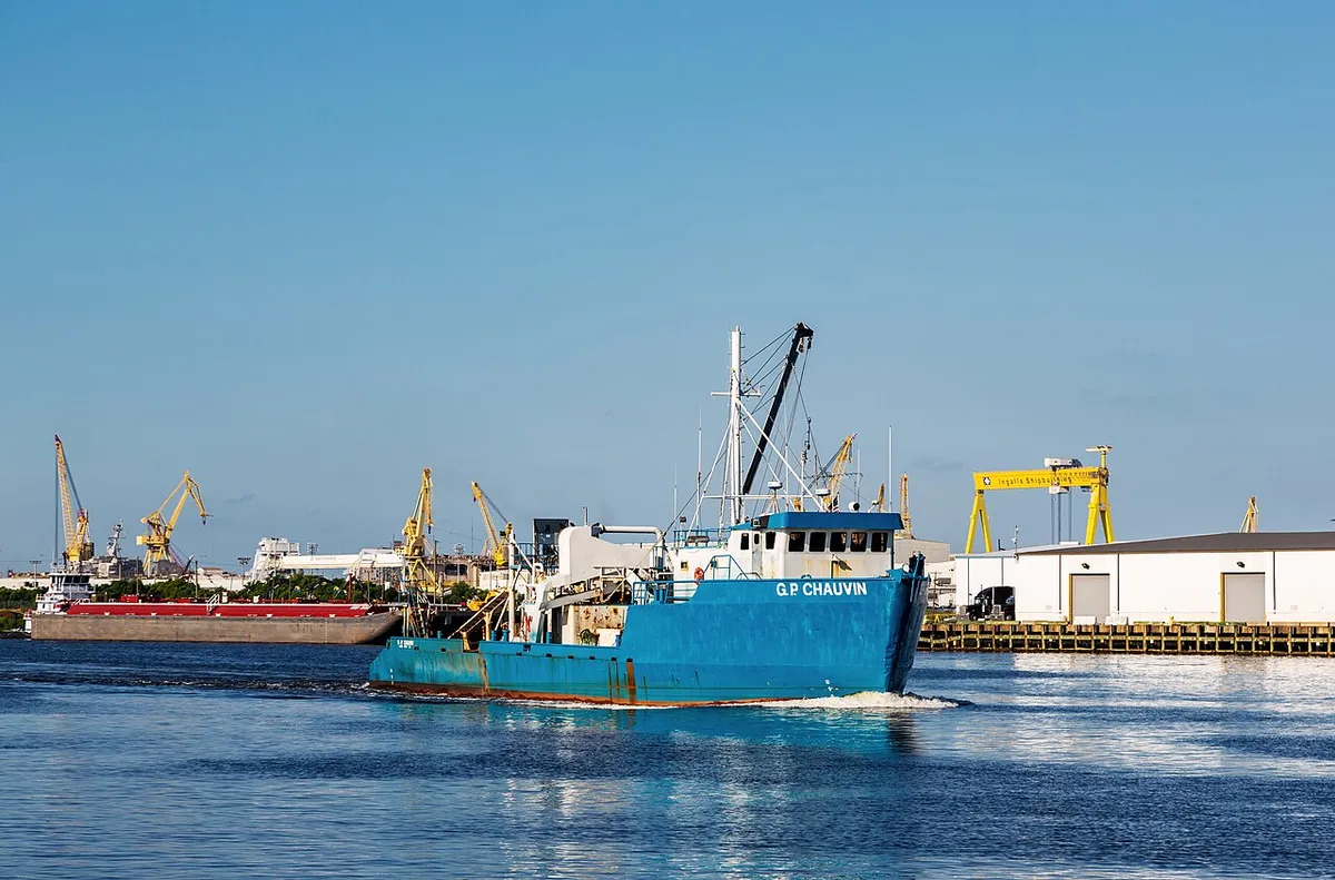 A blue-hulled work boat on a wide river in front of yellow shipyard cranes under a clear blue sky