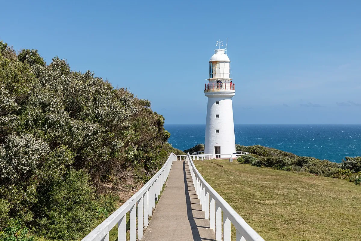 A white lighthouse on a grassy headland above a blue sea, with a walkway leading to it